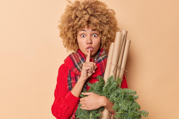 Mysterious woman makes silence gesture asks to be quiet presses index finger over lips tells secret of New Year traditions of celebration poses with spruce wreath and rolled paper poses indoor