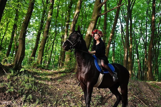 Mystical girl in wreath wear in black at horse in wood