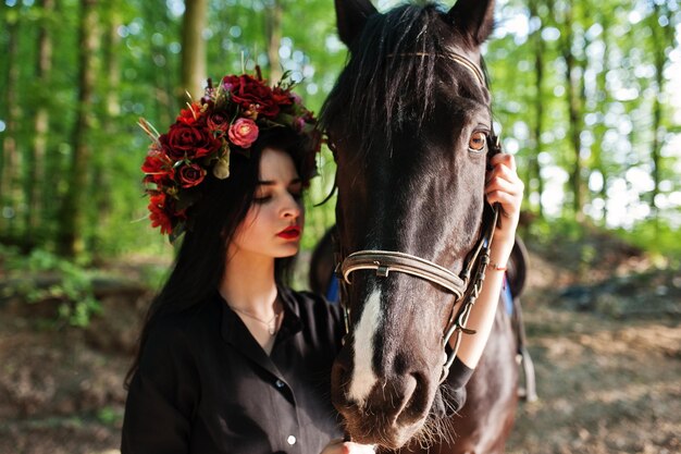 Mystical girl in wreath wear in black with horse in wood