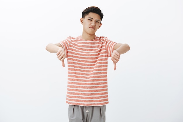Free photo nah giving negative feedback. displeased and dissatisfied attractive young asian male in striped t-shirt showing thumbs down raising head with disdain, being unimpressed