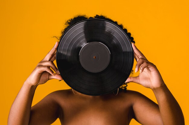 Naked African American young female covering face with vinyl plate in studio