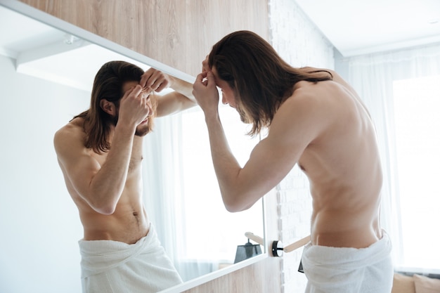 鏡の近くのにきびを絞るタオルで裸のひげを生やした若い男