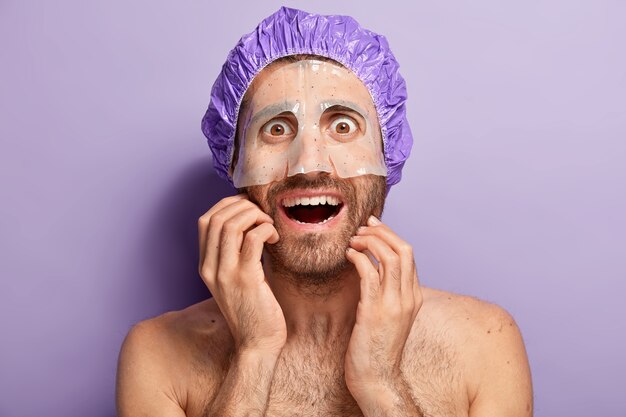 Naked happy man applies moisturize mask on face, wears violet shower cap, enjoys cosmetic skincare facial treatment, has brown bugged eyes, bare shoulders