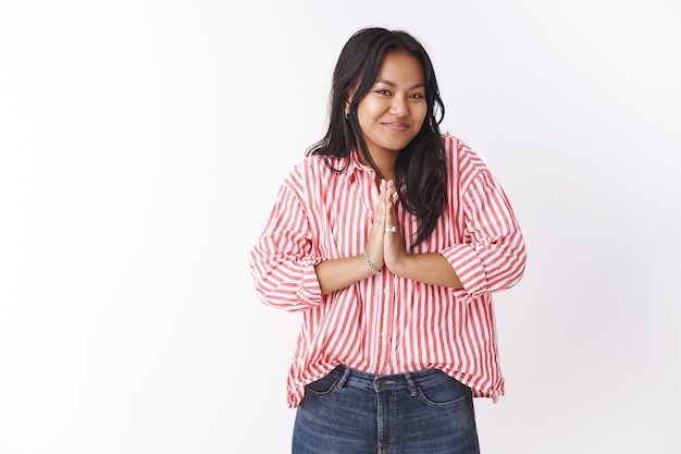 Namaste enjoy your stay. Portrait of cute pleasant and happy young nice vietnamese girl in striped blouse pressing palms together in asian greeting gesture smiling at camera upbeat over white wall