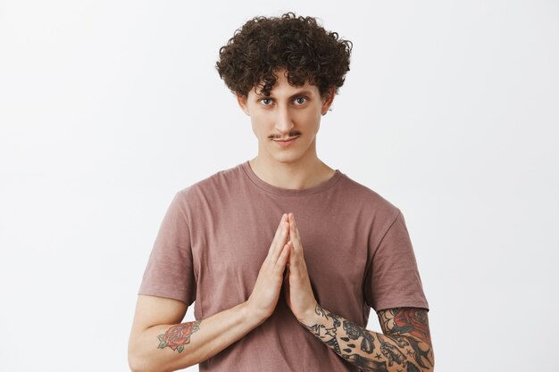 Namaste friends. Portrait of determined and mysterious attractive stylish city guy with moustache curly hairstyle and tattoos on arms holding palms together near body gazing over gray wall