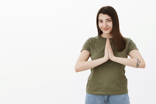 ナマステはあなたに平和をもたらします。灰色の壁に笑みを浮かべて祈るか仏のポーズでヤシの木を一緒に保持している入れ墨と茶色の髪のキュートでリラックスした見栄えの良い若い20代女性の肖像画