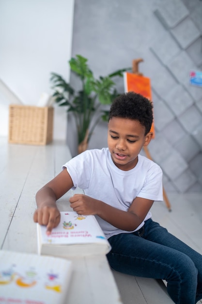 Name of book. Concentrated dark-skinned school-age boy in casual clothes sitting near window reading title of book touching page with his finger