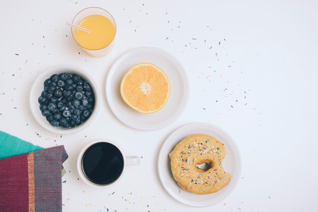 Napkin; blueberry; glass of juice; halved orange; coffee cup and donut on white backdrop
