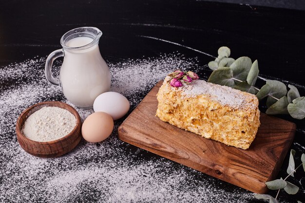 Napoleon cake decorated with dried flower seeds on black table with egg, milk and bowl of flour. 