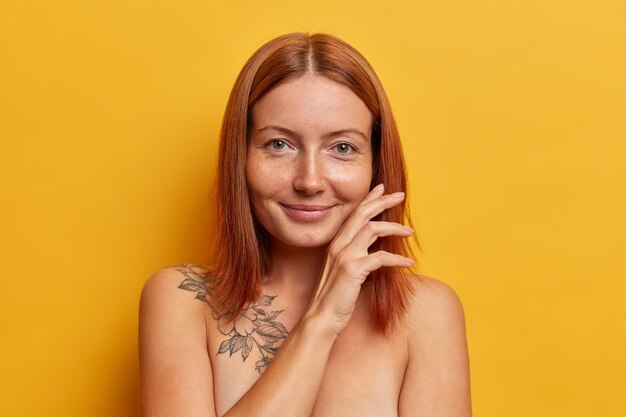 Natural beauty, purity and wellness concept. Satisfied redhead woman touches face gently, demonstrates her perfect smooth skin after spa procedure, stands bare shoulders against yellow wall