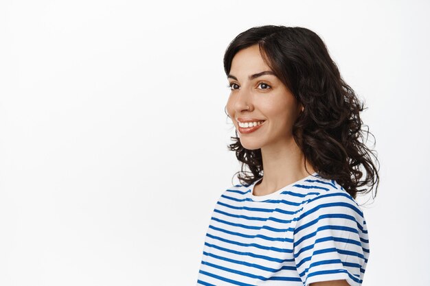 Natural brunette girl with pierced nose and curly hairstyle, looking aside at empty space on white, wearing striped t-shirt