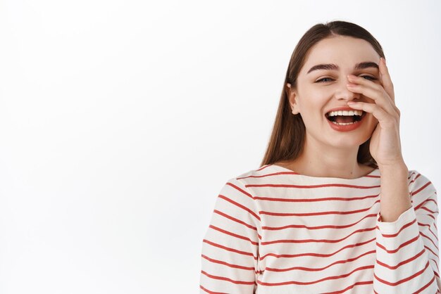 Natural candid girl laughing and looking happy smiling with white perfect teeth holding hand on clean healthy face standing over studio background