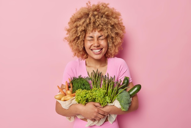 Natural food concept Joyful woman with curly hair keeps eyes closed smiles toothily poses with freshly picked potatoes carrot asparagus and lettuce isolated over pink background Eco products