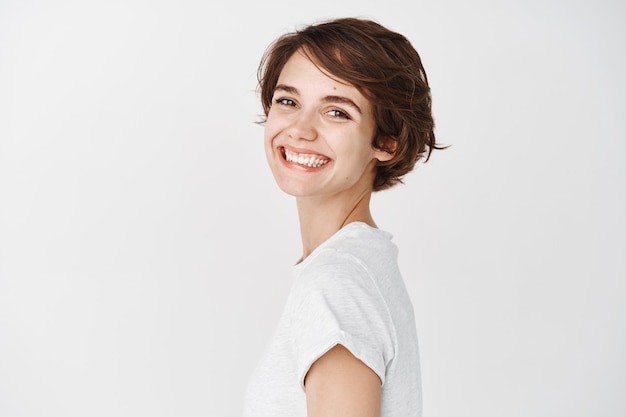 Giovane donna naturale con un sorriso felice, gira la testa e sembra allegra, in piedi contro il muro bianco in maglietta