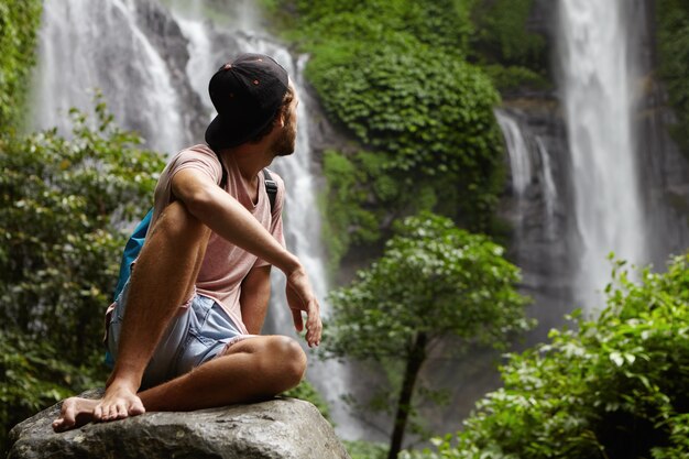 Nature, wildlife and travel concept. Young barefooted hiker wearing snapback sitting on big stone and enjoying beautiful view around him. Hipster relaxing deep in rainforest