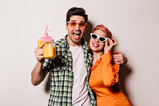 Непослушный мальчик и девочка в стильных нарядах и солнцезащитных очках обнимаются, улыбаются и позируют с оранжевым коктейлем на белом пространстве.