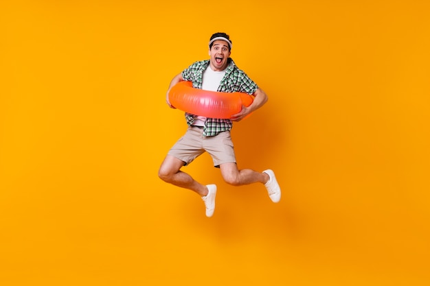 Naughty brunet male dressed in plaid shirt and beige shorts jumping with inflatable circle on isolated space.