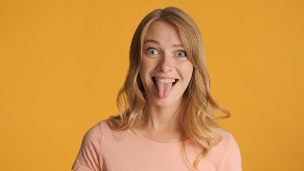 장난 꾸러기 재미있는 금발 소녀 노란색 배경 위에 카메라에 혀를 내밀고 표현 주위를 속이고