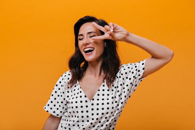 Naughty girl laughs and shows peace sign on orange background. Charming brunette in white polka-dot clothes is smiling and having fun.