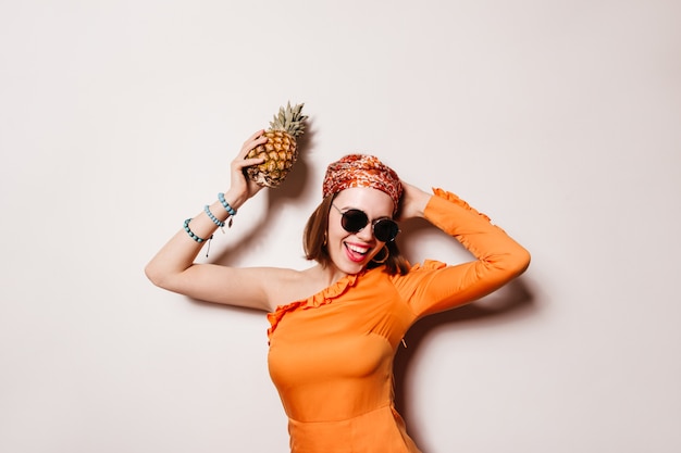 Naughty girl in orange dress, sunglasses and with blue bracelet on her arm is dancing and holding pineapple.