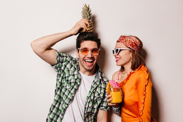 Naughty man in green shirt holds pineapple on his head and looks into camera with smile. Lady in orange blouse and glasses holding cocktail.