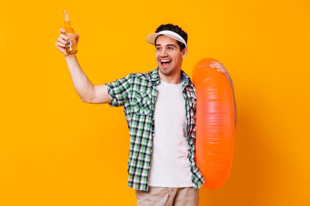 Naughty man picks up bottle of beer, laughs and holds inflatable circle on isolated space.