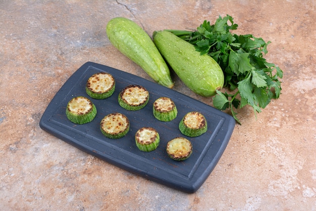 Navy tray of fried zucchini slices next to a bundle of parsley and a whole raw zucchini on marble surface