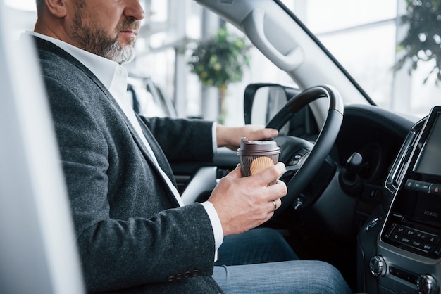 Need to charge with some coffee. Side view of senior businessman in official clothes inside of modern car