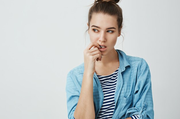 Need to create some plan. Portrait of thoughtful attractive caucasian girl with bun hairstyle looking aside while biting fingernail, thinking about something, being nervous .