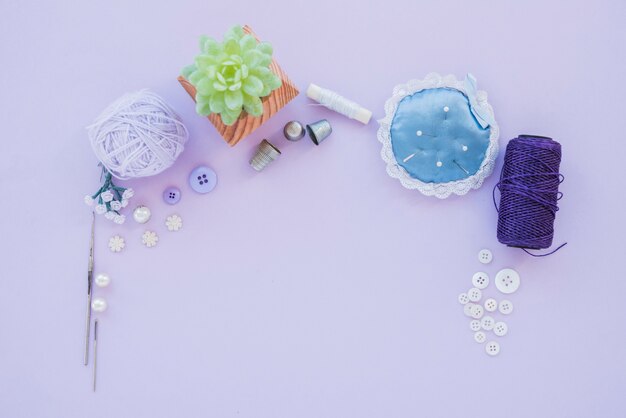 Needles with pincushion; thimble; wool ball; beads; button and yarn spool on purple backdrop