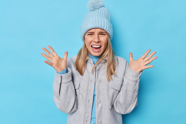 Negative emotions concept. Emotional woman screams loudly with anger keeps palms raised reacts emotionally on something negative wears knitted hat and grey jacket isolated over blue background