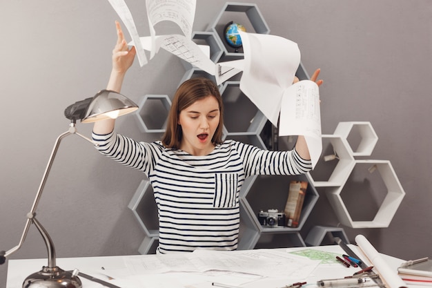 Free photo negative emotions. portrait of angry young good-looking female architect throwing papers away, being angry with bad result of work after lot of tries.