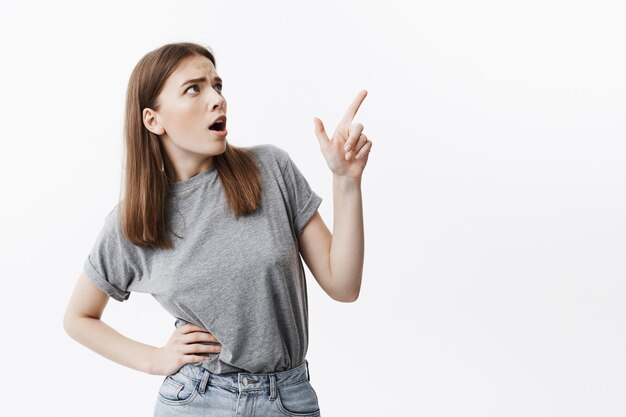 Negative emotions. Portrait of expressive young good-looking student girl with dark hair in grey clothes looking aside with opened mouth and mean expression, pointing on white wall with finger. Copy