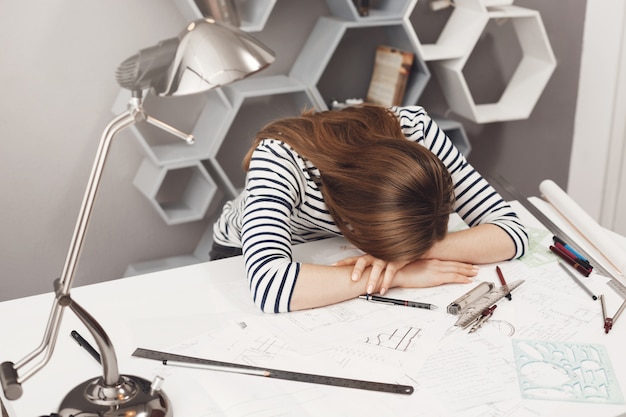 Free photo negative emotions. portrait of young fashionable female freelance architect lying on hands at table, being tired after working too much, dreaming of sleep