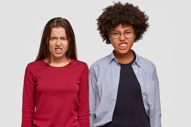 Negative facial expressions concept. Displeased multiethnic women frown face in irritation, clench teeth, look with outraged expression, annoyed with bad words or comments, isolated over white wall