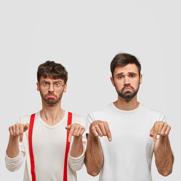 Negative facial expressions and feeling concept. Upset two men with gloomy expressions point down, frown faces with displeasure, dislike something, pose over white wall with blank space