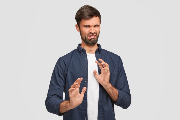 Negative facial expressions and people concept. Unhappy displeased young male frowns face with discontent look, feels aversion towards something, gestures indoor, stands against white wall