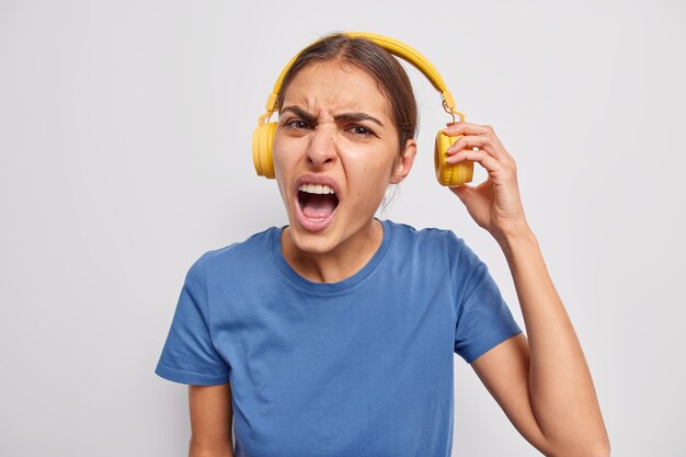 Негативная разочарованная европейская женщина снимает наушники, слушает музыку с громким звуком, снимает наушники, чтобы избежать шума в ушах, носит повседневную синюю футболку, изолированную над серой стеной