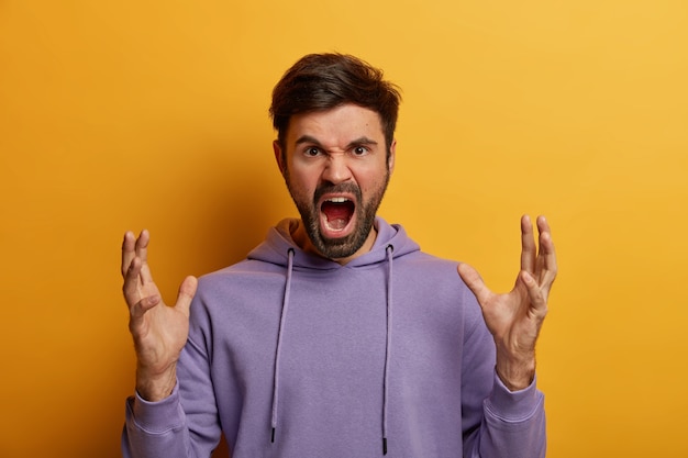 Negative human emotions and feelings. Angry annoyed bearded adult man screams loudly, expresses irritation, gestures angrily, keeps palms raised, reproaches someone and has quarrel, wears hoodie