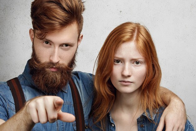 Negative human face expressions and emotions. Angry Caucasian man with fuzzy beard hugging his shy and upset girlfriend, pointing his index finger  as sign of reproach.