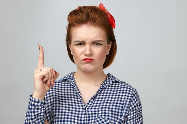Negative human facial expressions, emotions and reaction. Emotional unhappy young glamorous female dressed in checkered shirt frowning and pouting, having upset grumpy look, raising index finger