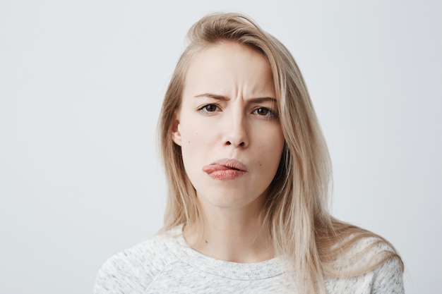 Negative human reaction, feelings and attitude. Close-up portrait of disgusted squeamish blonde woman in casual wear grimacing, sticking out tongue, feeling nauseous because of bad smell or stink