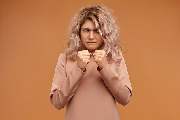 Negativity, aggression and rage concept. Funny emotional young Caucasian woman frowning, holding clenched fists in front of her, ready to punch enemy