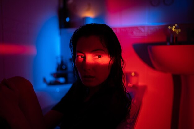 Неоновый портрет молодой депрессивной азиатки с полосой красного света на лице, принимающей ванну в одежде