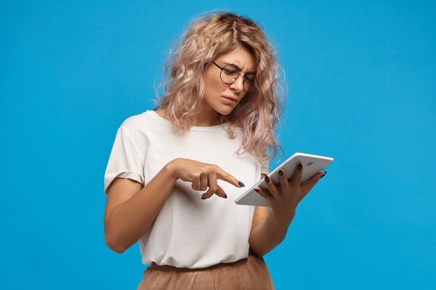 터치 패드 디지털 태블릿에서 인터넷을 서핑하는 둥근 안경에 괴상한 진지한 힙 스터 소녀, 소셜 네트워크를 통해 사진 스크롤