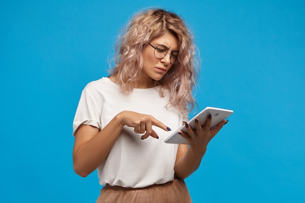 Nerdy serious hipster girl in round spectacles surfing internet on touch pad digital tablet, scrolling pics via social networks