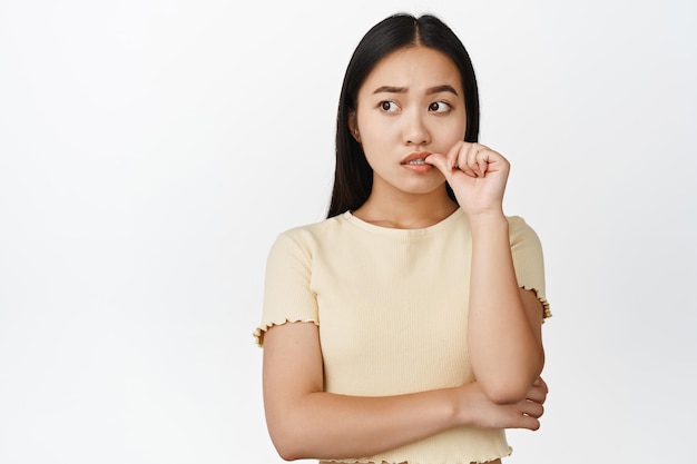 Нервная азиатская девушка кусает палец и смотрит в сторону, думая и волнуясь, стоя в желтой футболке на белом фоне