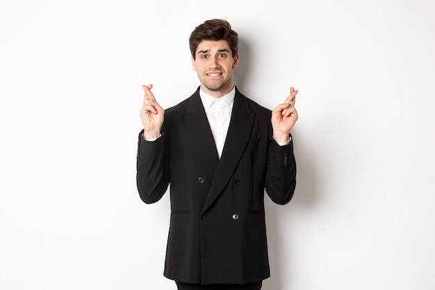 Nervous businessman in black suit crossing fingers, biting lip and making a wish, waiting for news, standing over white background hopeful.