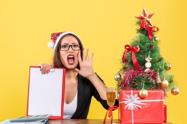 Nervous charming lady in suit with santa claus hat and eyeglasses showing document in the office 