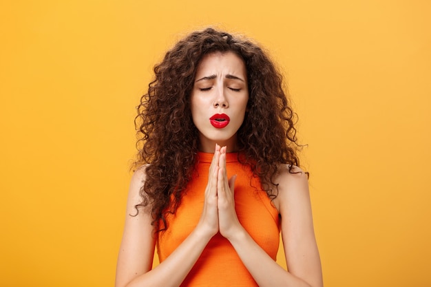 Nervous and concerned woman with curly hairstyle feeling hopeful praying with closed eyes and frowne...